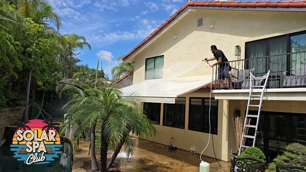 Technician softwashing the stucco wall of a two-story home with a tile roof from a second-floor balcony